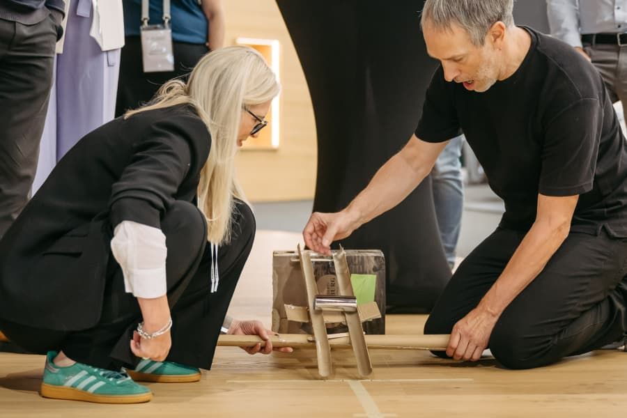 Heidi Blaschek kniet neben einem weiteren Workshop-Teilnehmer am Boden - sie bauen gemeinsam eine einfache Konstruktion aus Papprollen, Karton und Holzstäben.